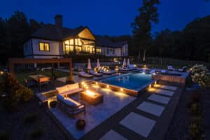 Nighttime aerial view of a luxury estate featuring a glowing rectangular pool, an outdoor lounge with a lit fire pit, a dining pergola, and a modern home with brightly lit windows.