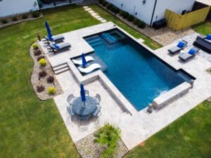 Bird's-eye view of a geometric swimming pool with a dark blue finish, featuring a built-in spa, a tanning ledge with white loungers, and a light gray travertine stone deck with outdoor dining and seating areas.