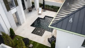 High-angle view of a modern T-shaped swimming pool with a dark bottom finish, surrounded by a light-colored travertine stone patio and a white luxury home with a black metal roof.