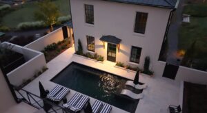 A high-angle view of a small rectangular plunge pool in a private walled courtyard, featuring black and white striped lounge chairs and a modern white two-story home.