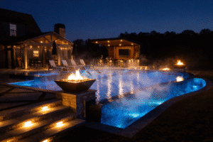 A luxury inground swimming pool at night in Georgia featuring a dark blue finish, infinity edge, and integrated stone fire bowls. Steam rises from the water’s surface, demonstrating active circulation and pool freeze protection protocols during a winter deep freeze.